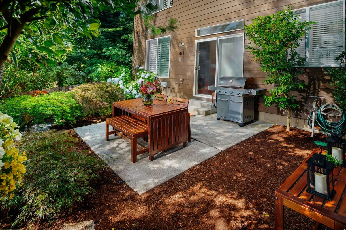 A casual and welcoming backyard patio scene with a wooden dining table and benches, a grill ready for summer cookouts, and vibrant flower arrangements. The backdrop features rich greenery, emphasizing the harmonious indoor-outdoor transition.