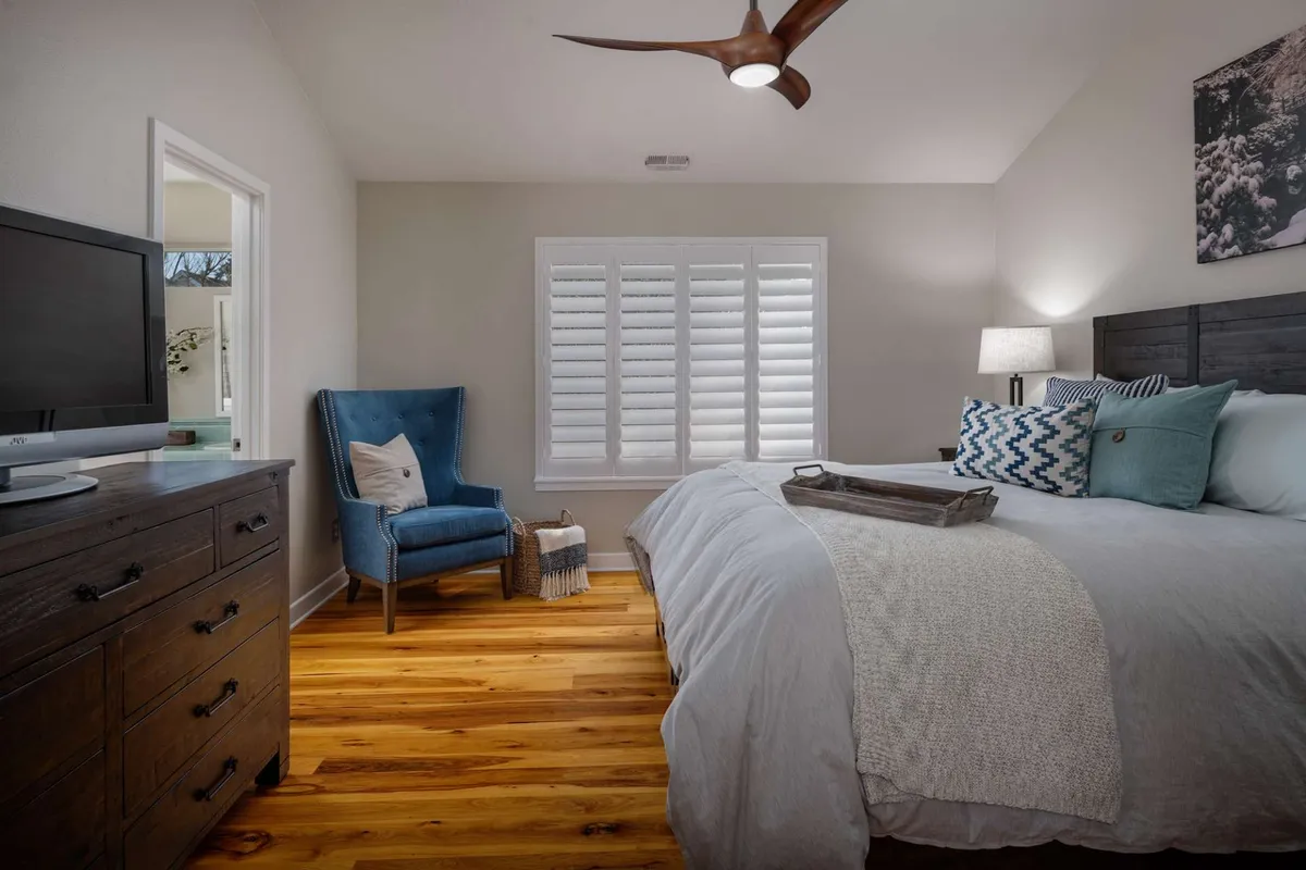 A serene bedroom with a textured blanket draped across the bed, a blue accent chair in the corner, a wooden dresser, and a ceiling fan overhead. Natural light brightens the space, complementing the calm and comfortable setting.