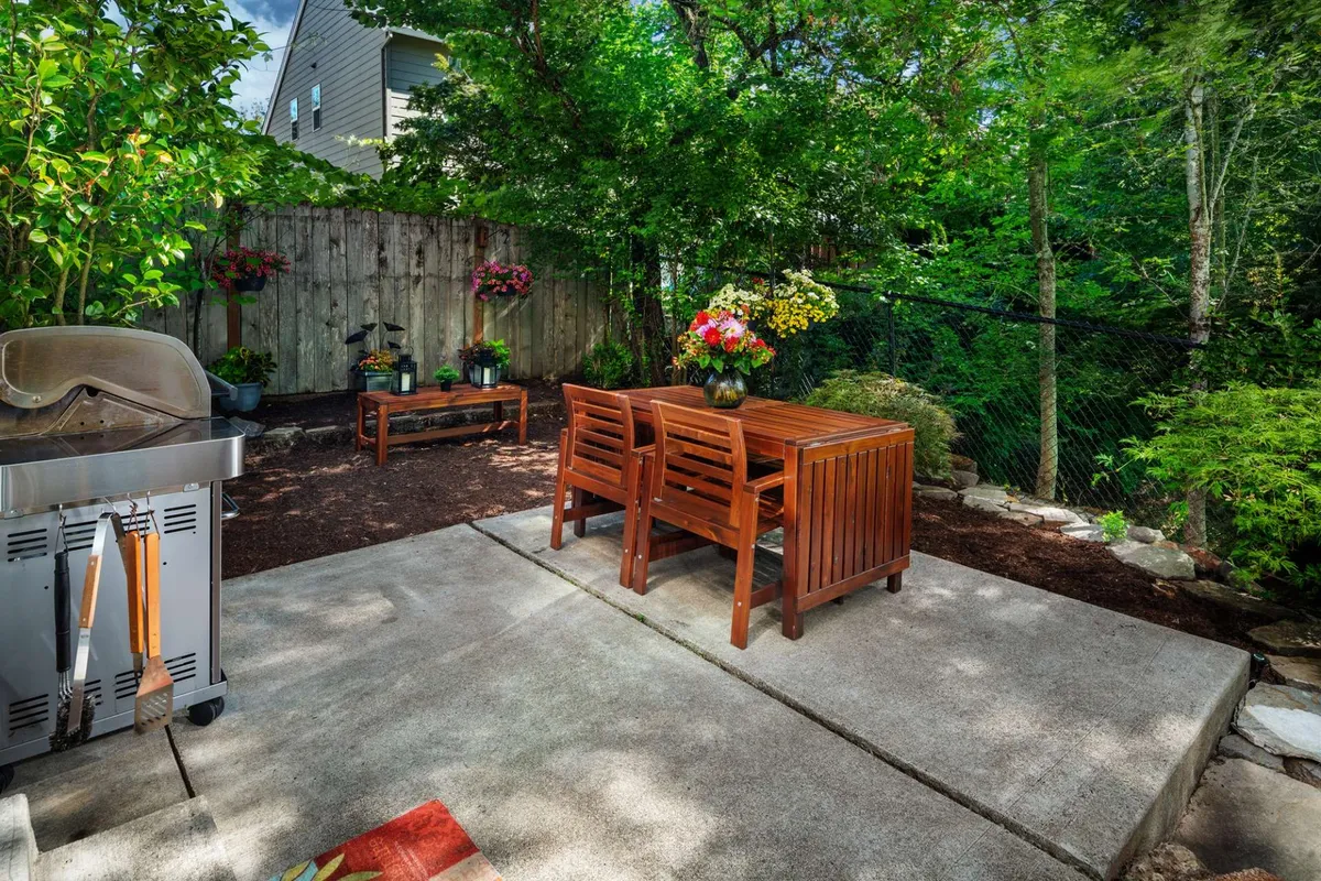 A relaxed backyard view showcasing a wooden dining table, an outdoor grill, and flower pots surrounded by greenery. The inviting setup is perfect for social gatherings, gardening, or simply unwinding outdoors.