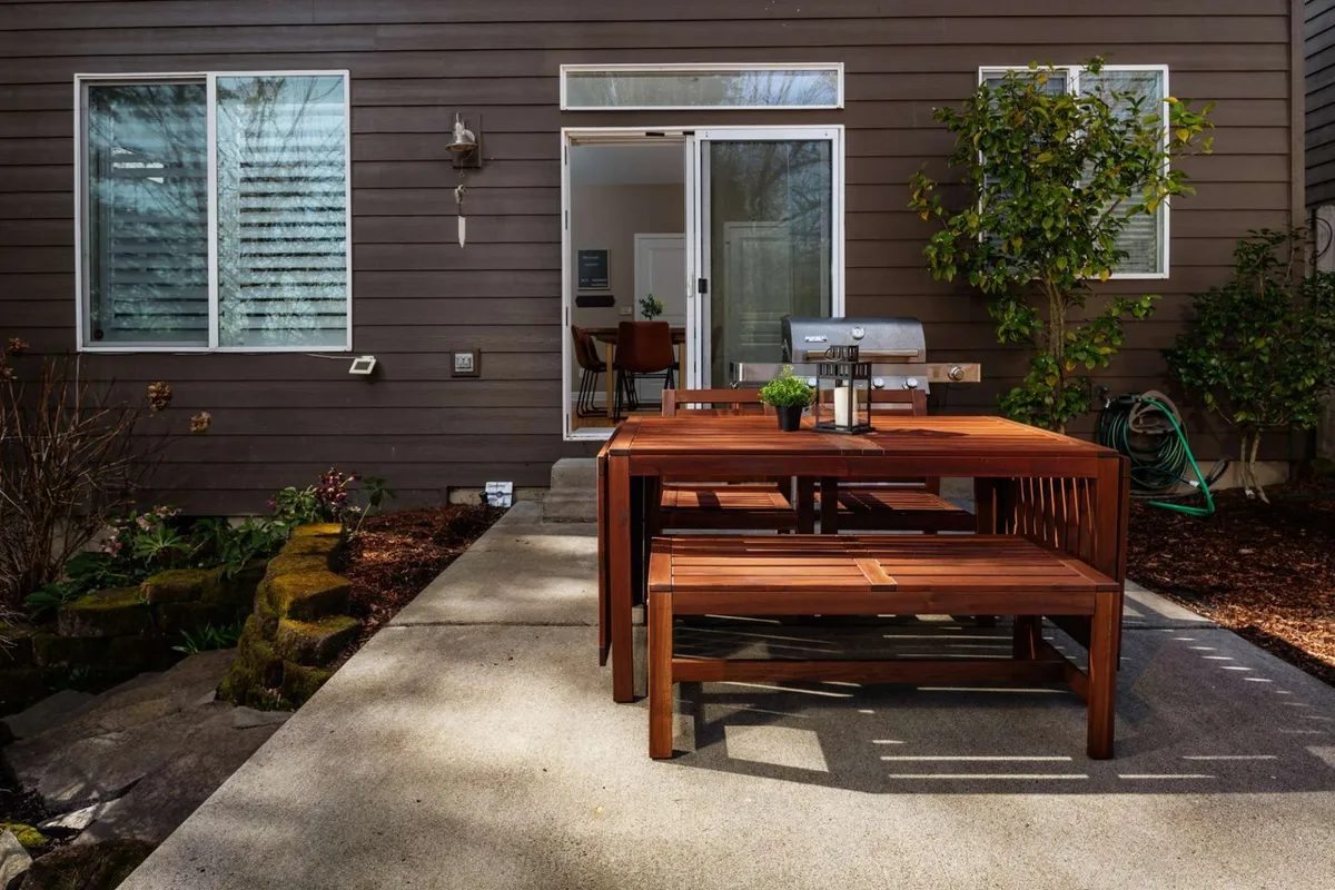 This exterior image showcases a cozy outdoor dining area with a wooden table, grill, and abundant green plants. The relaxed mood highlights opportunities for unwinding after work, socializing, or enjoying a peaceful meal outdoors—defining the appeal of nearby amenities for off-the-clock relaxation.