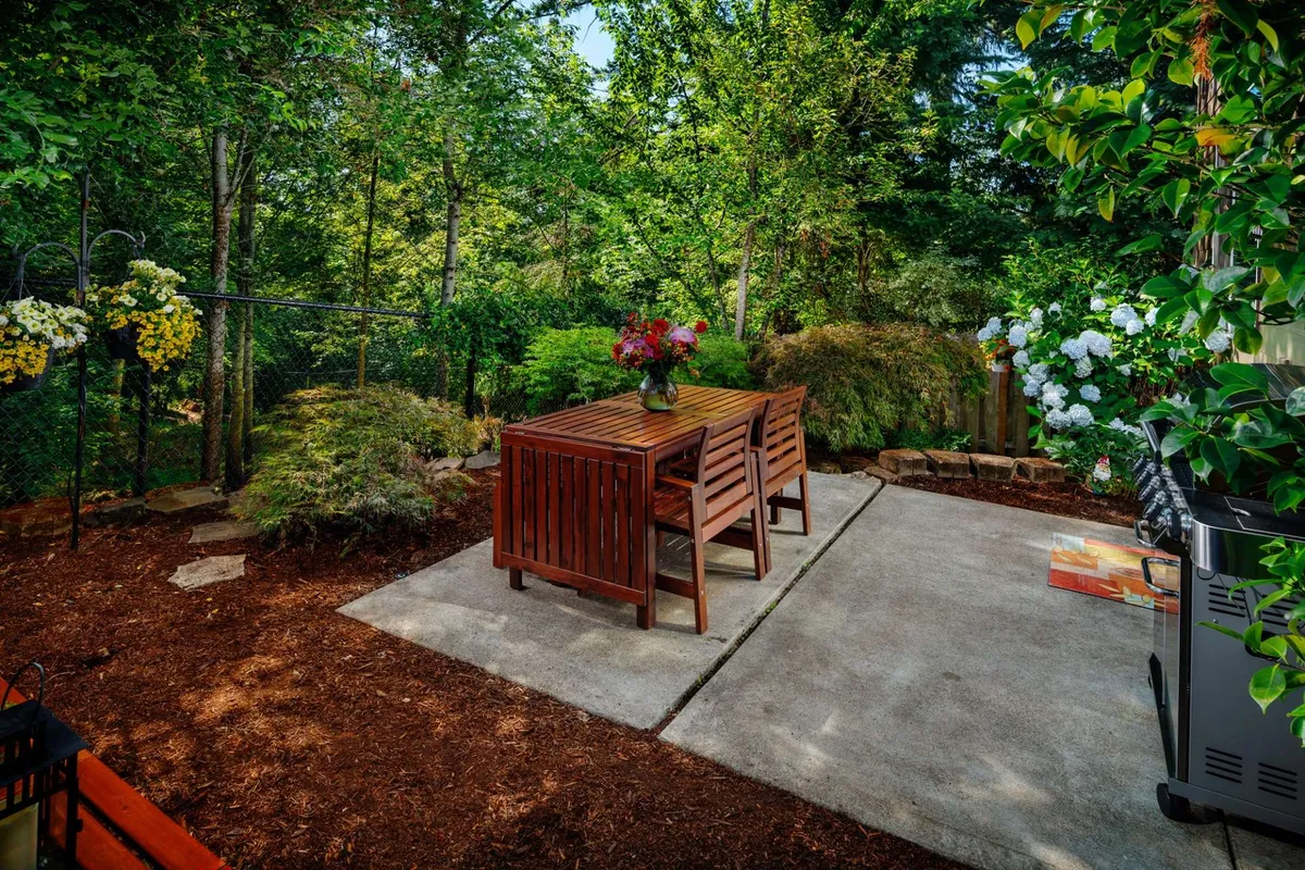 A serene private patio featuring a wooden dining set with a vase of fresh flowers, surrounded by lush greenery and a stone pathway leading away from the seating area. The tranquil atmosphere is ideal for outdoor dining, morning coffee, or relaxing amid nature.