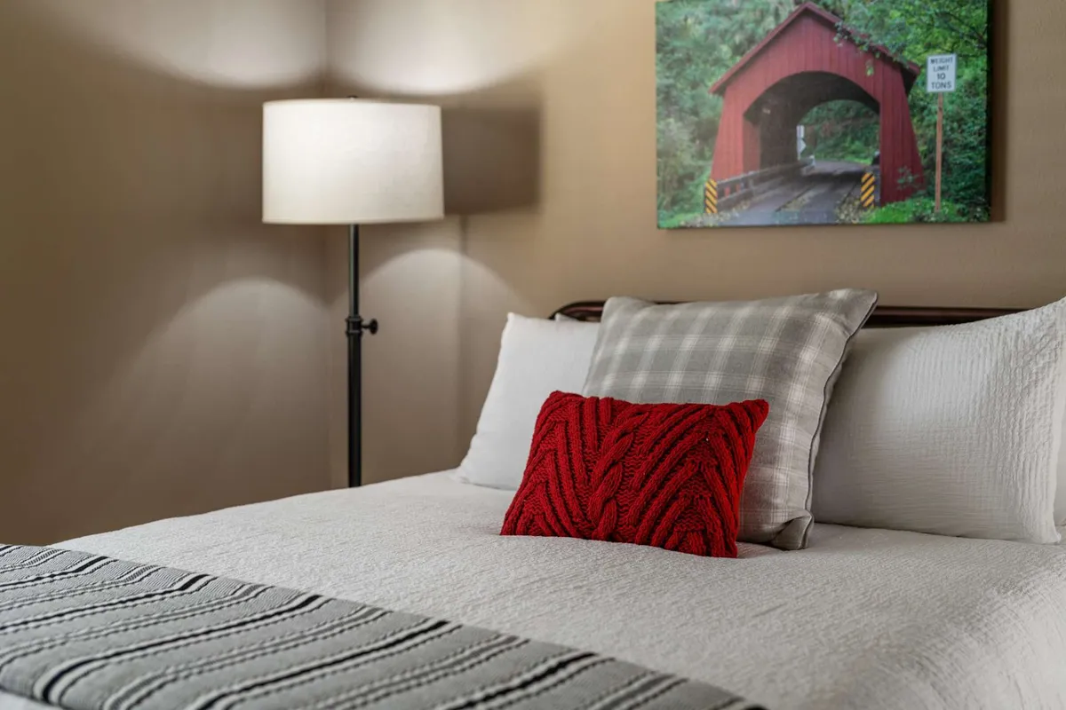 A cozy bedroom featuring a bed with decorative pillows, a glowing table lamp, and framed artwork of a covered bridge on the wall. Warm, ambient lighting and soft bedding create a welcoming environment ideal for restful sleep and relaxation.